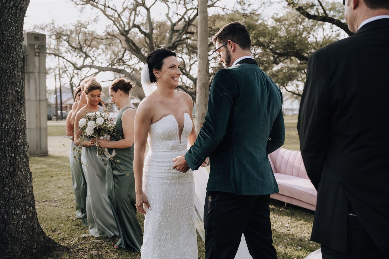 Elegant outdoor wedding ceremony with attendees in formal attire under tree canopy.
