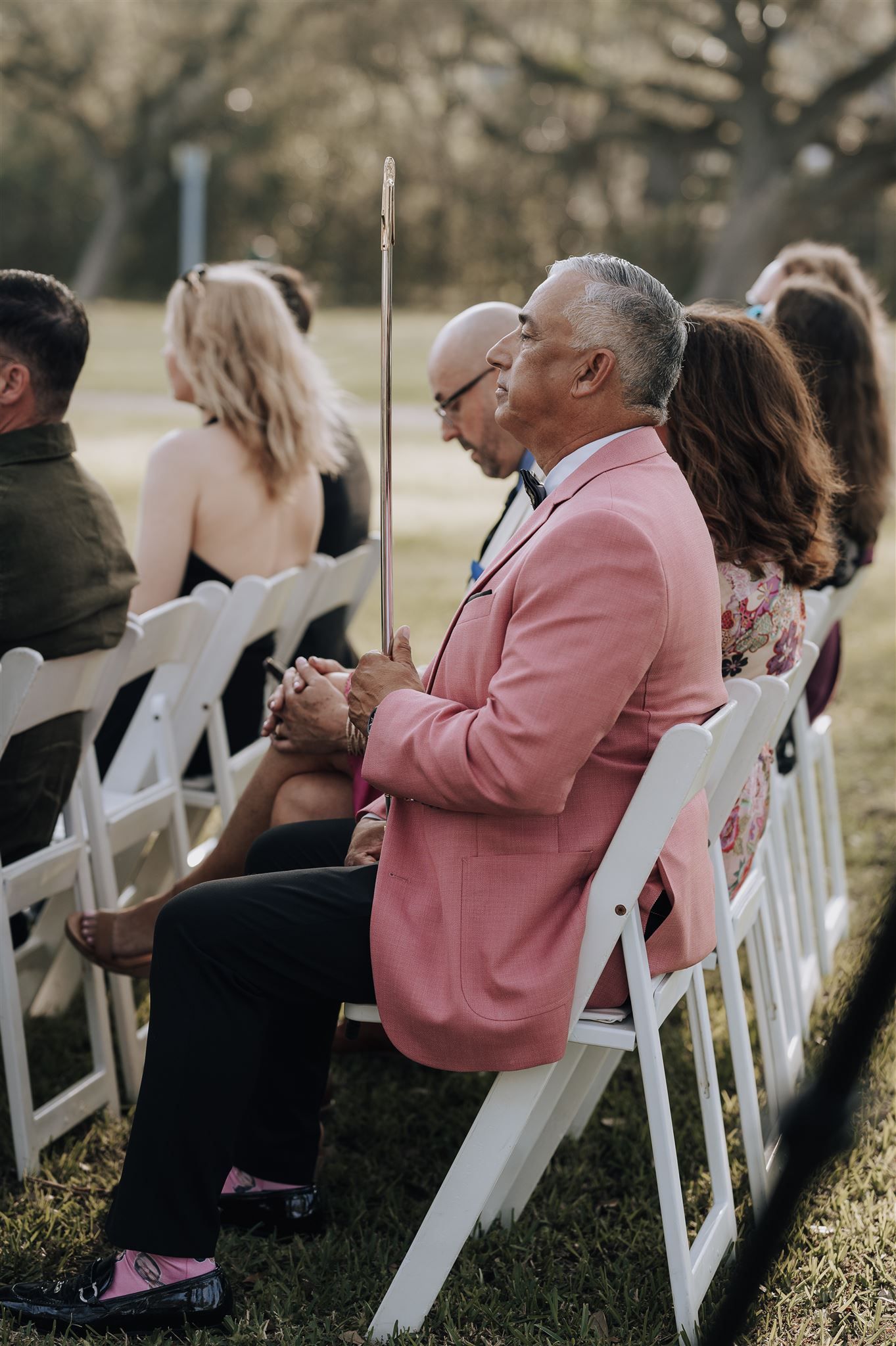 Wedding guest in pink blazer seated on white folding chair during outdoor ceremony.