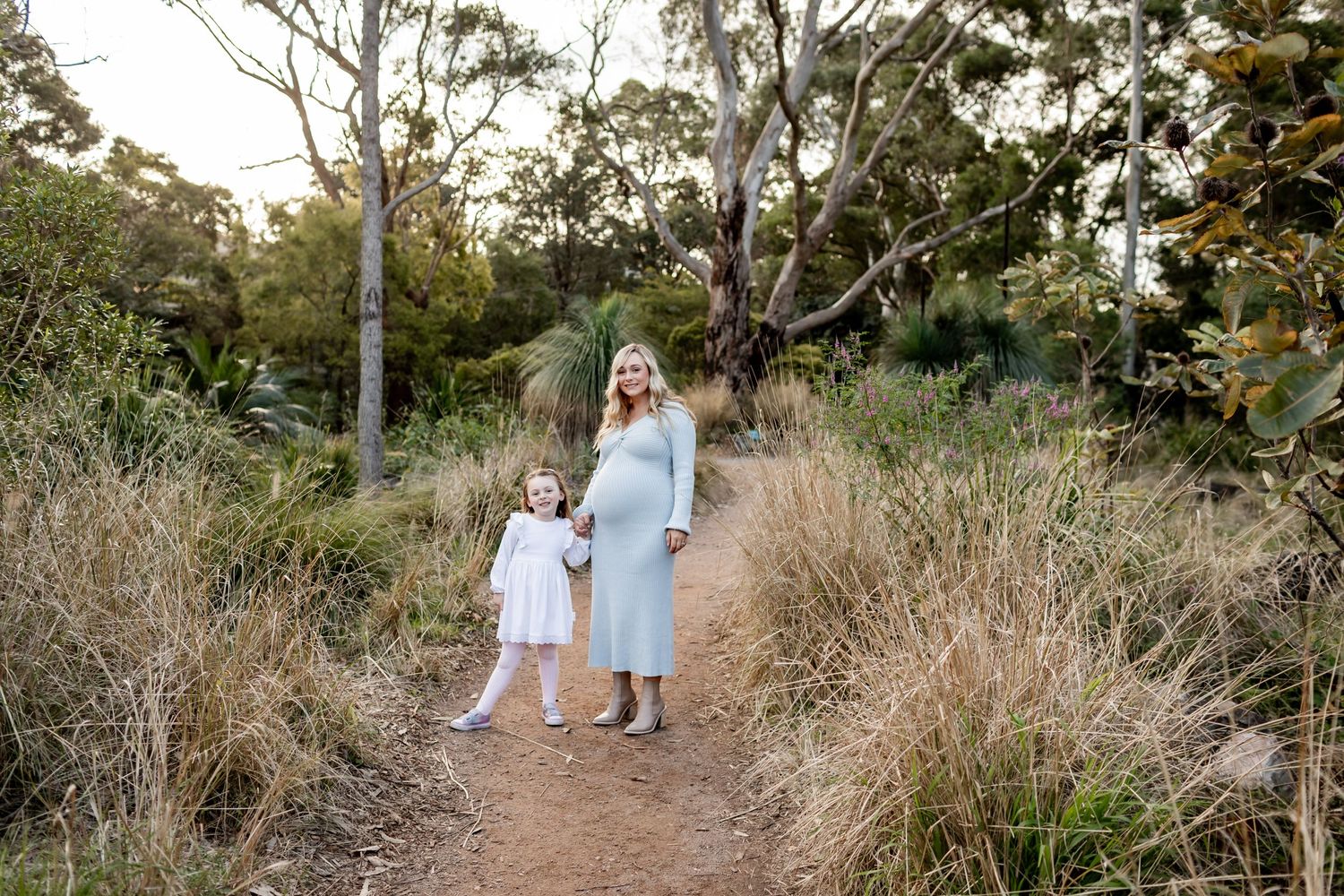 Two people in light-colored outfits walk together on a winding dirt path through natural bushland.