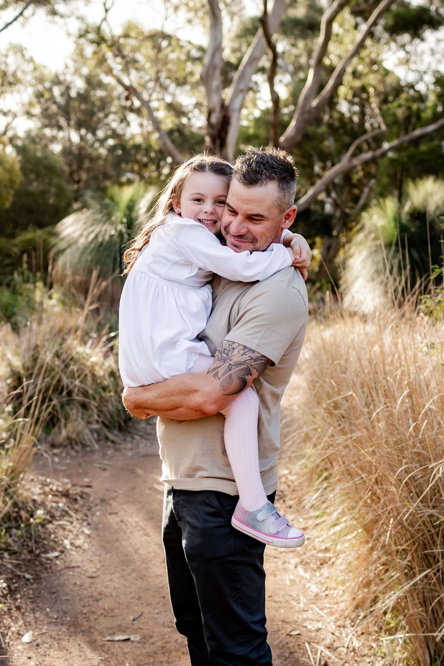 Father holds daughter in white dress on nature trail while sharing joyful hug.