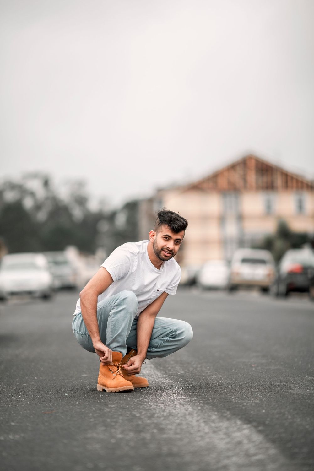 A person in casual white t-shirt and light jeans crouches down to tie their tan work boots in a parking lot.