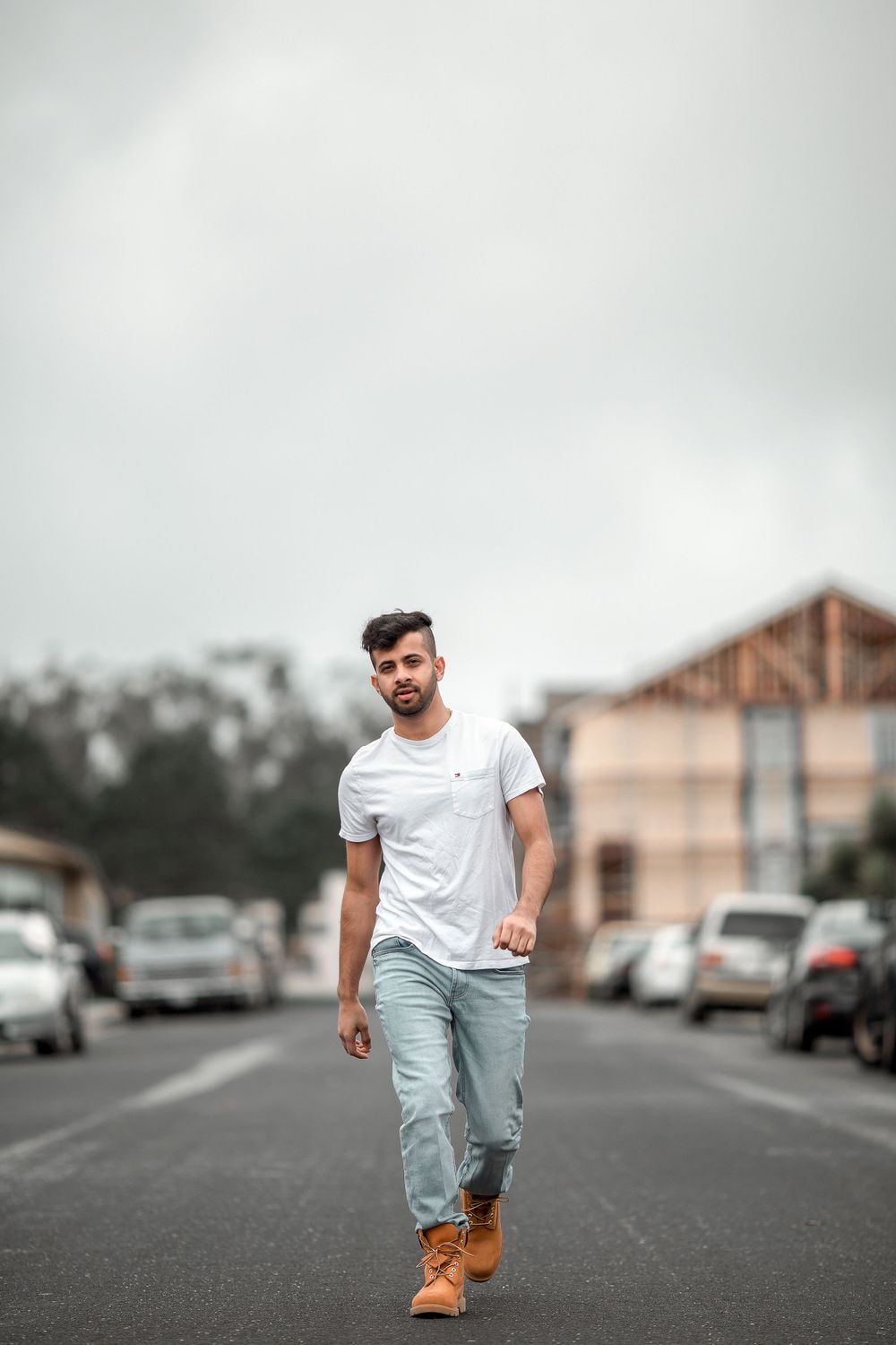 Person in white t-shirt and light jeans walking confidently down an urban street with parked cars and buildings in background.