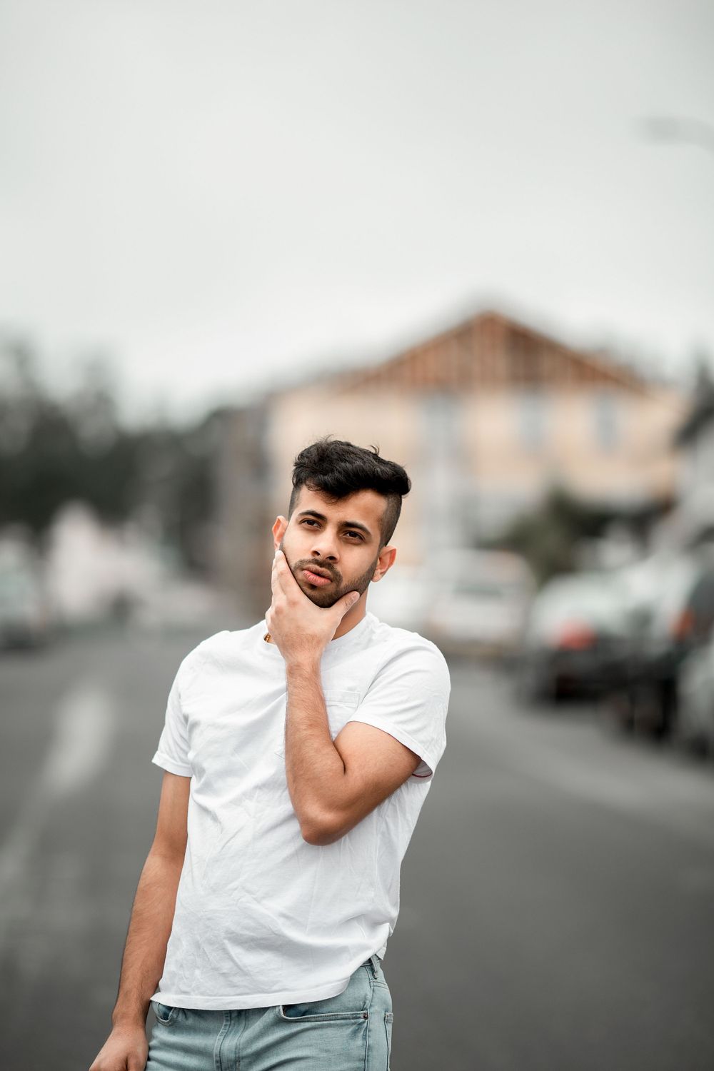Person in white t-shirt and gray pants standing thoughtfully on suburban street with houses in background.