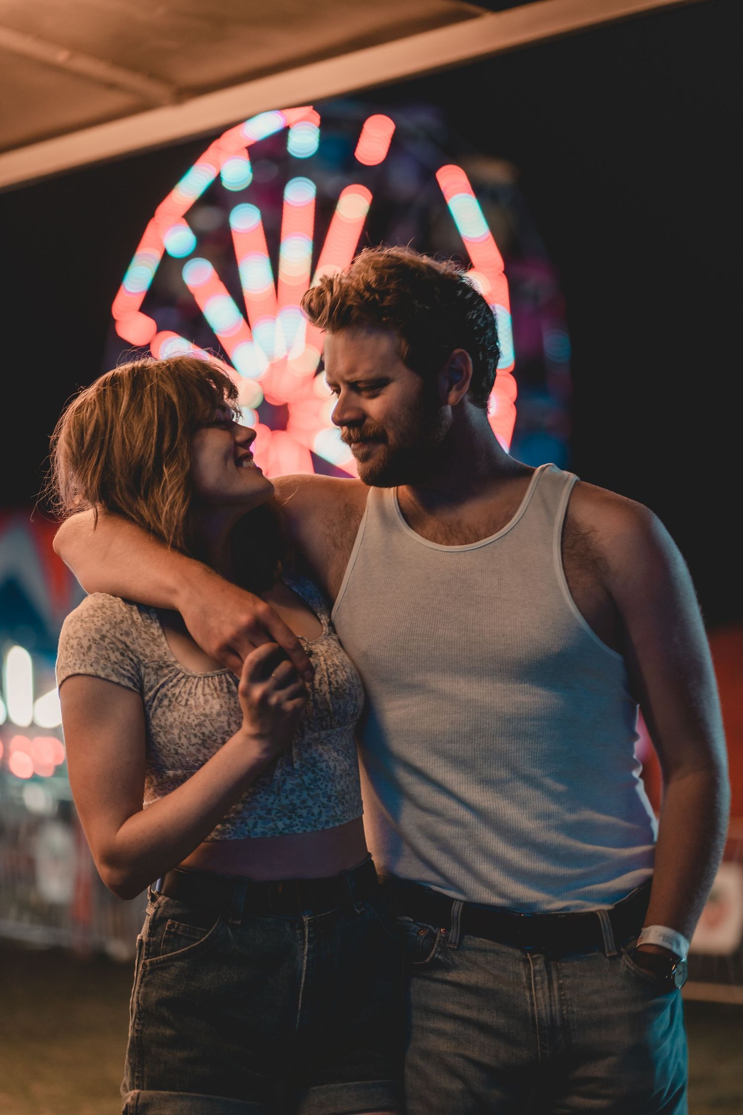 Tender embrace between couple beneath colorful carnival lights at dusk.