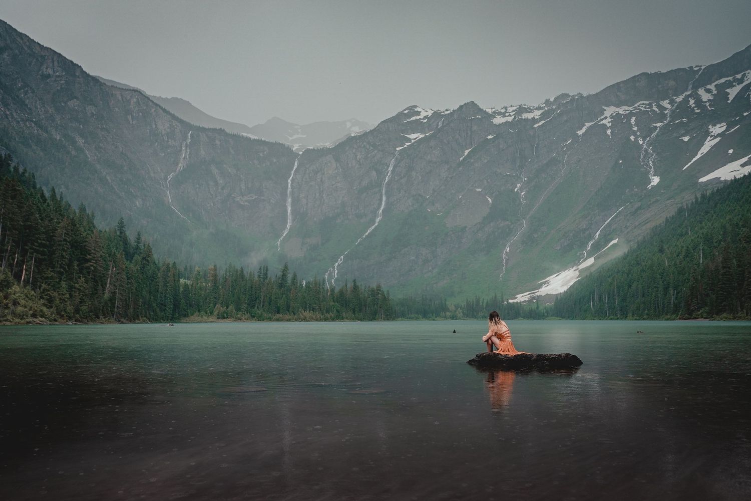 A lone figure sits in a boat on a still mountain lake with towering peaks and waterfalls in the background.
