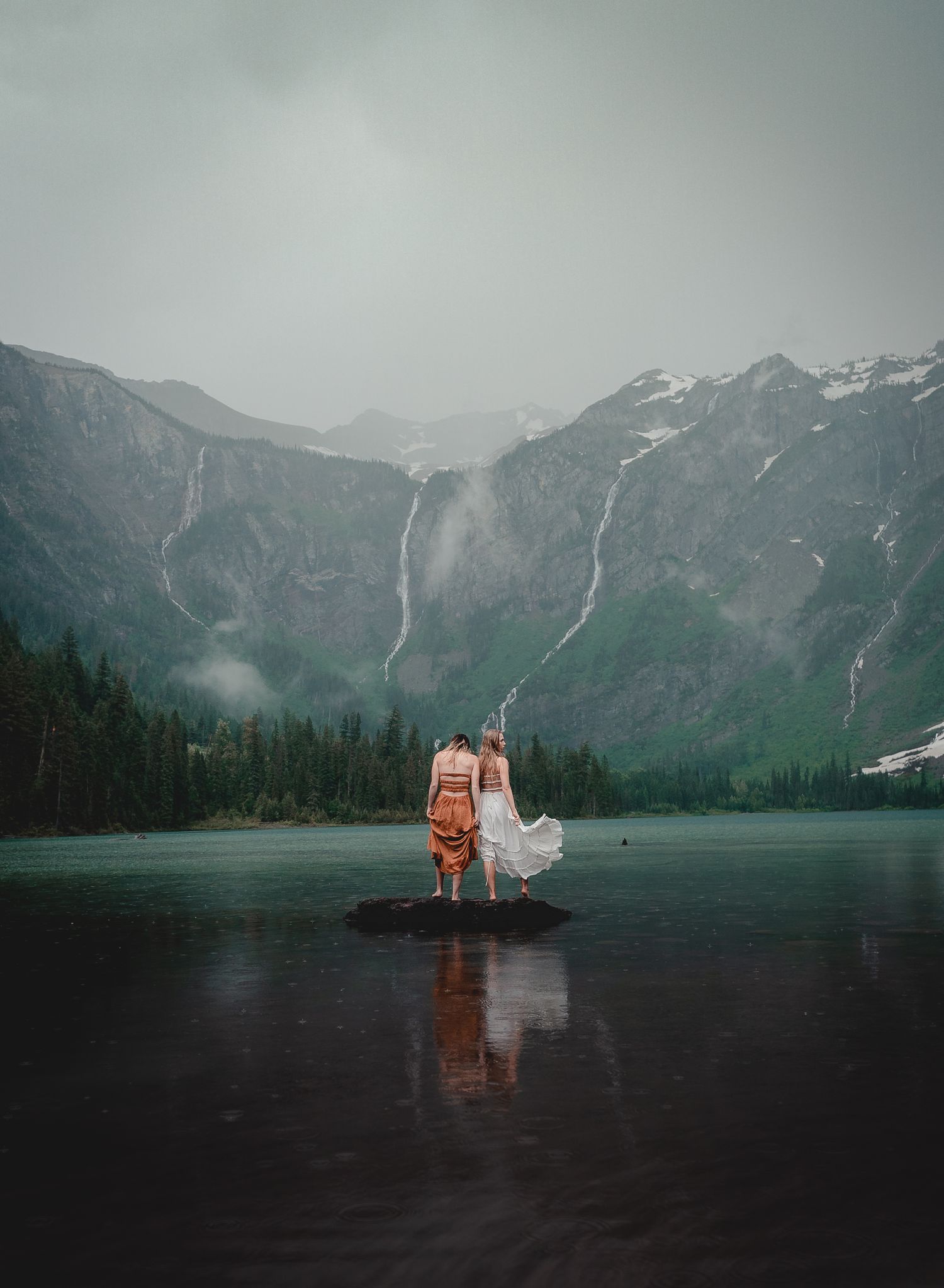 A couple stands on a frozen lake surrounded by snow-capped mountains and waterfalls in a dramatic landscape.