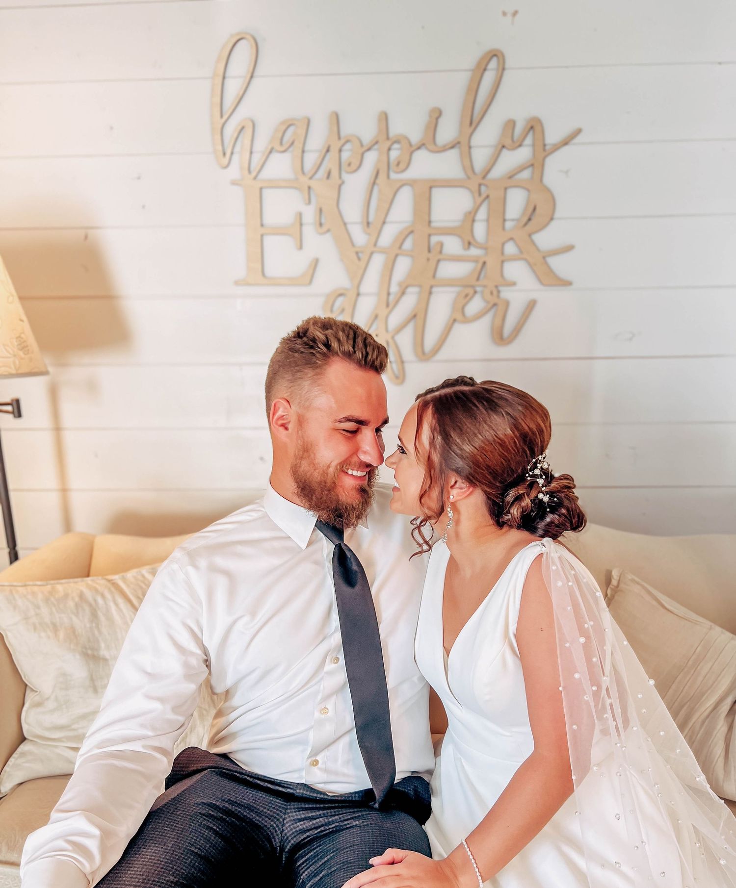 A romantic couple poses under a 'Happily Ever After' sign in elegant wedding attire.