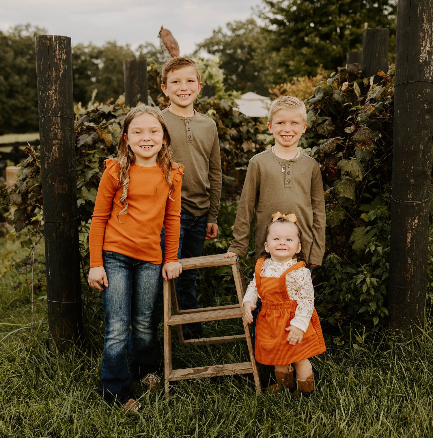 Four children stand together by wooden posts in autumn garden setting.