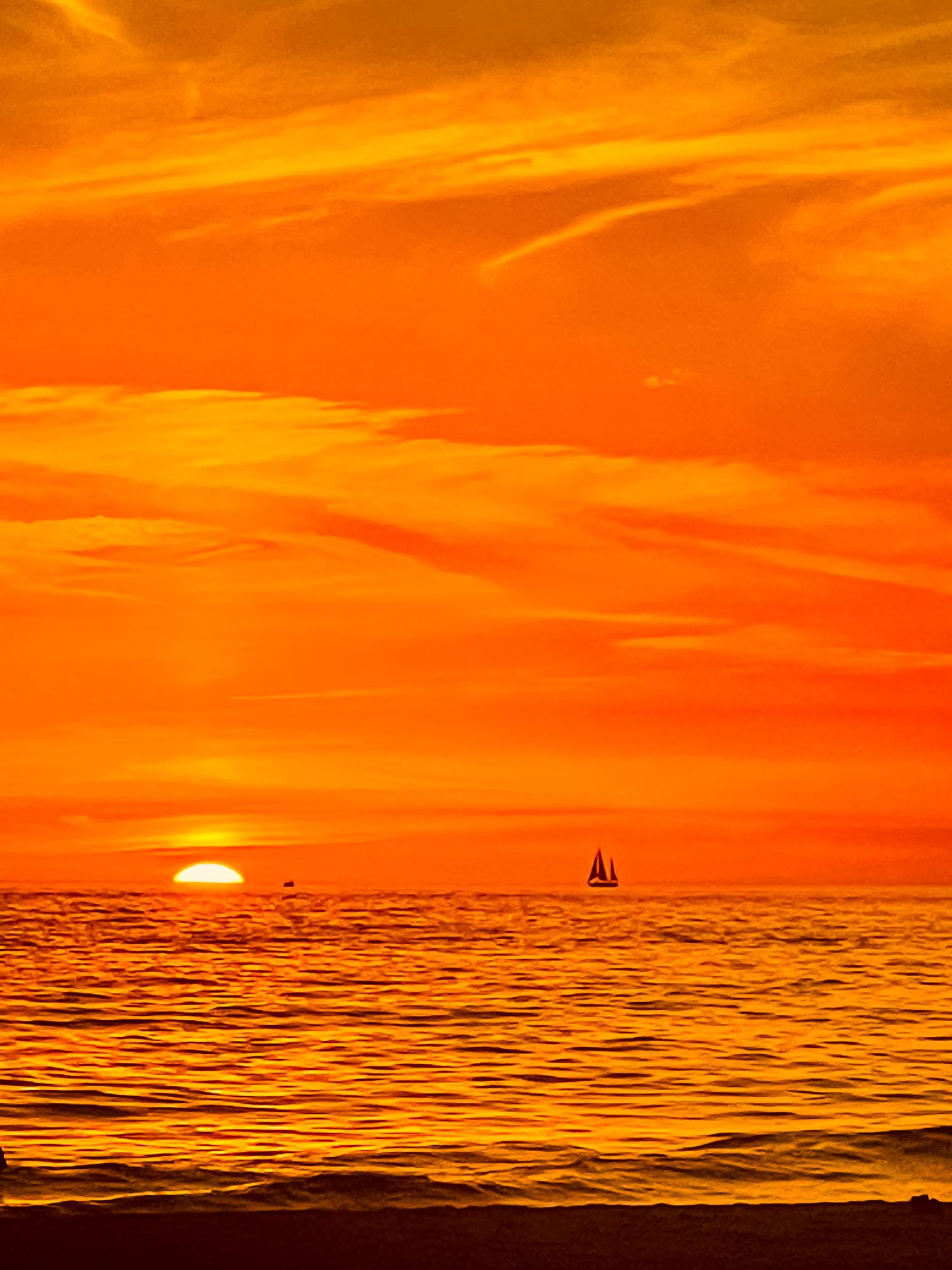 Sailboat silhouetted against brilliant orange sunset sky over ocean horizon.