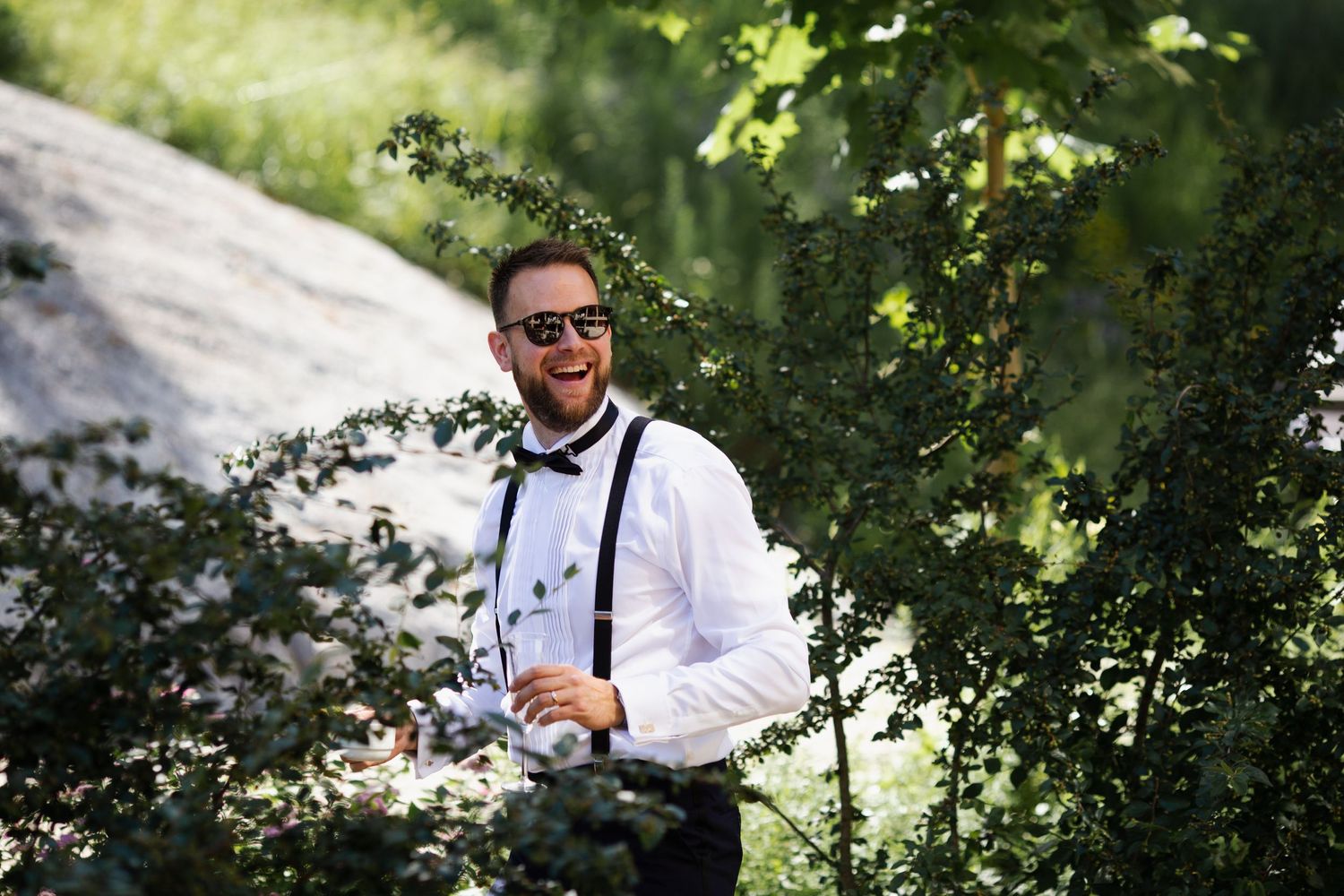 Professional photographer in formal attire with suspenders and sunglasses poses in a natural outdoor setting.