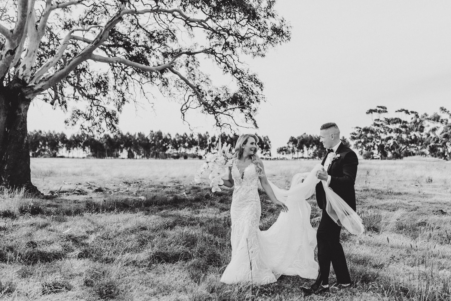 Black and white wedding portrait captured in open field under large tree.