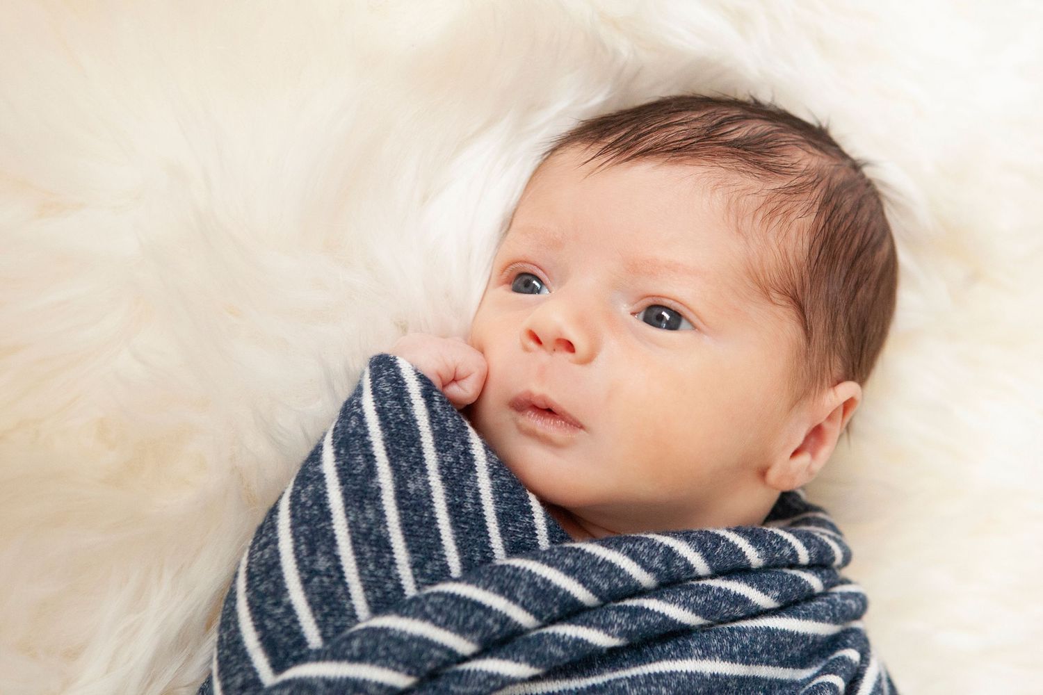 A baby in striped clothing gazes at the camera while lying on soft white fur.