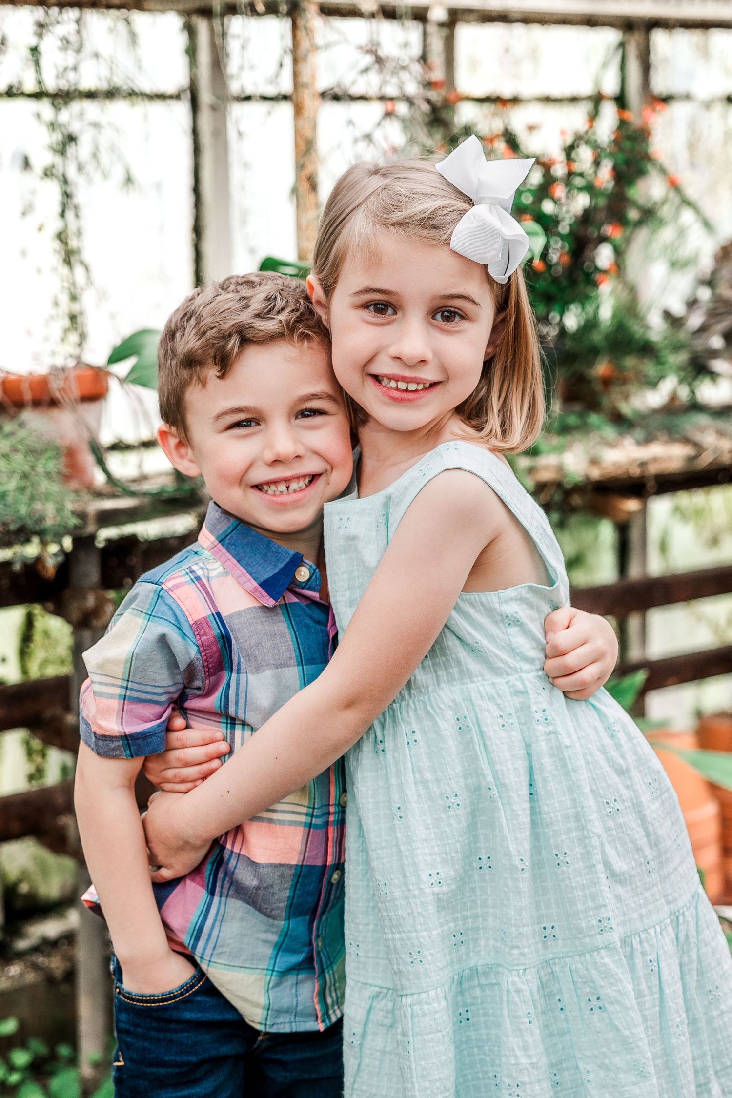 Two figures embrace while posing in mint green and plaid outfits in a greenhouse.