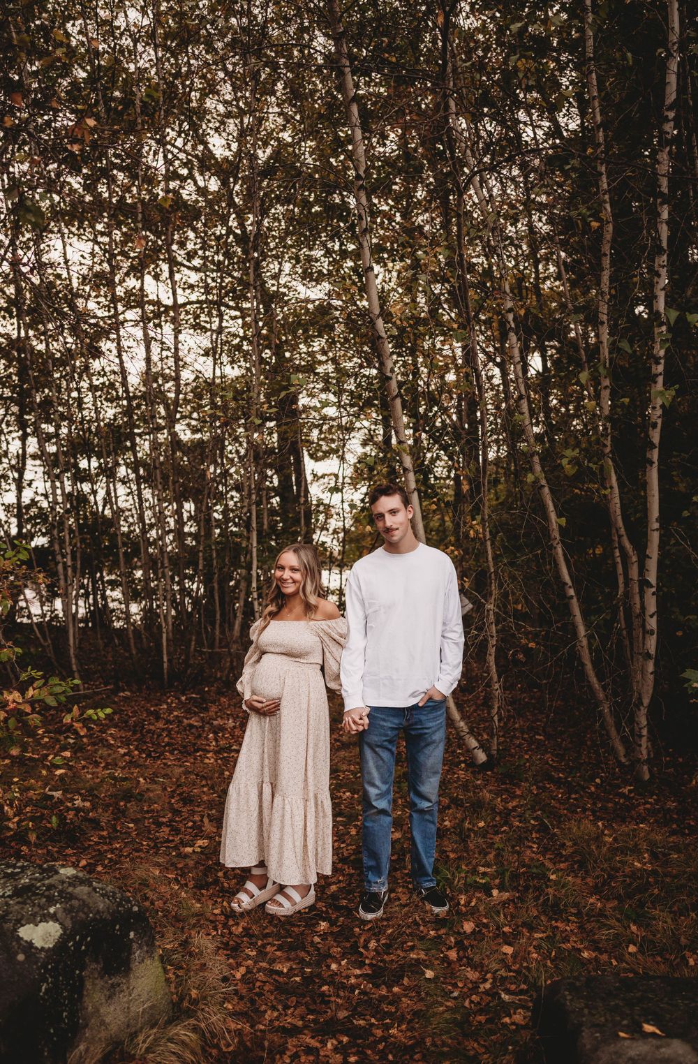 A couple stands together on fallen leaves in an autumn forest setting.