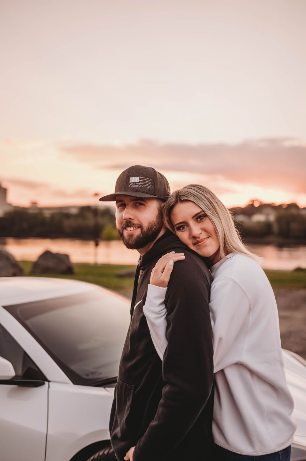 A couple leans against a white car during sunset with warm golden light.