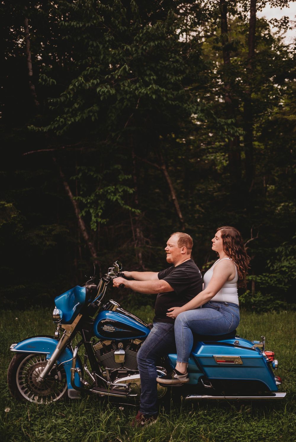 A couple sits together on a bright blue motorcycle in a serene forest setting.