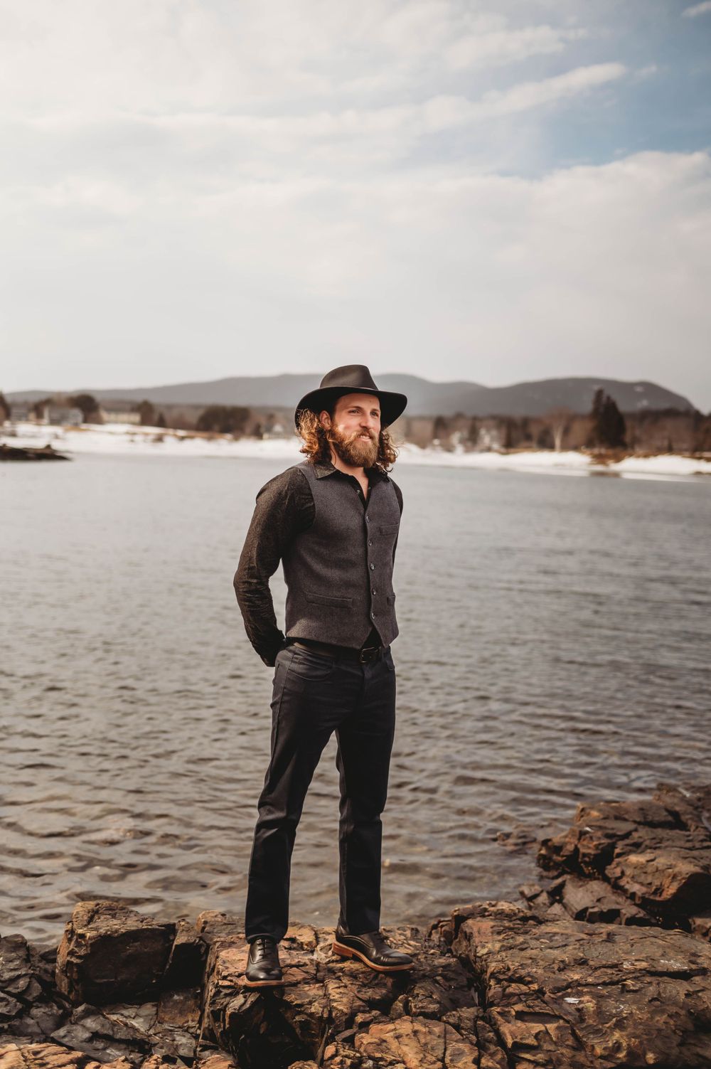 Person in dark clothing and hat stands on rocky shore overlooking winter lake landscape.
