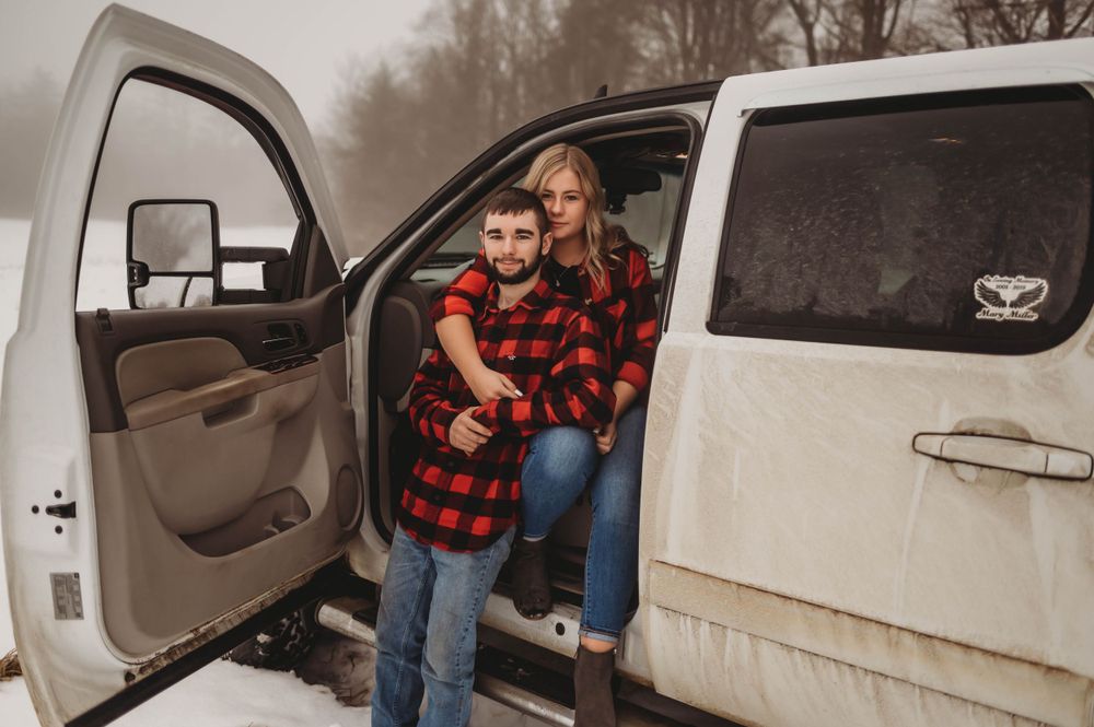 A couple wearing red buffalo plaid flannel shirts stands by an open car door on a foggy winter day.