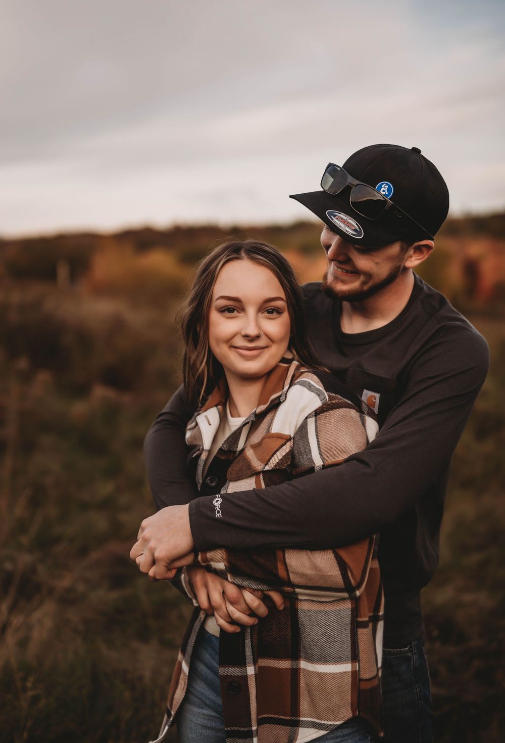 A couple embraces in casual fall attire against an autumn landscape.