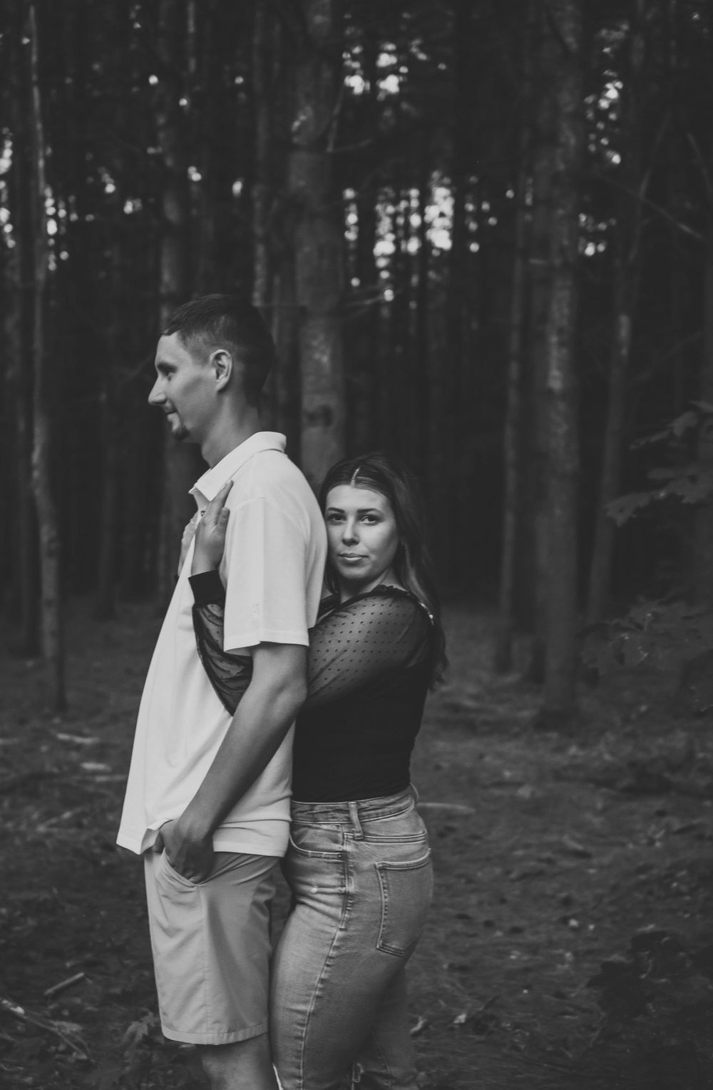 Black and white portrait of a couple posed back-to-back in a dark forest setting.
