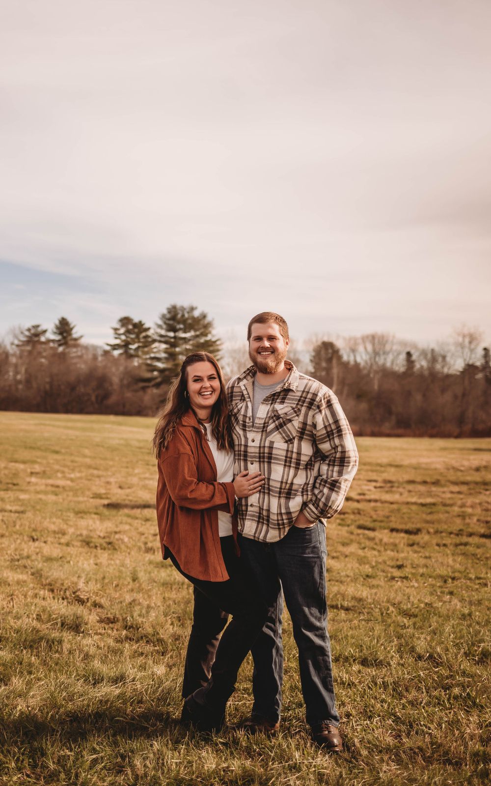 Couple poses together in rustic plaid shirts against autumn field backdrop.