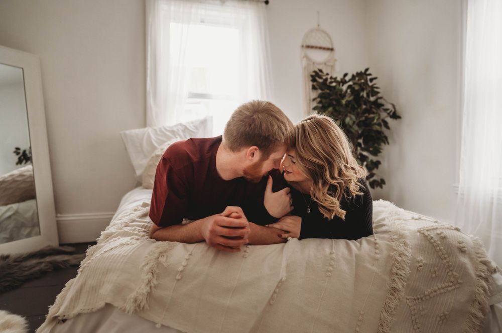 A couple lying together on white bedding shares intimate moment in bright bedroom setting.