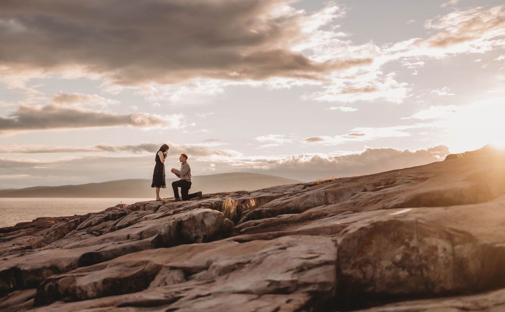 A silhouetted couple stands on rocky cliffs during golden hour with mountains and water in the distance.