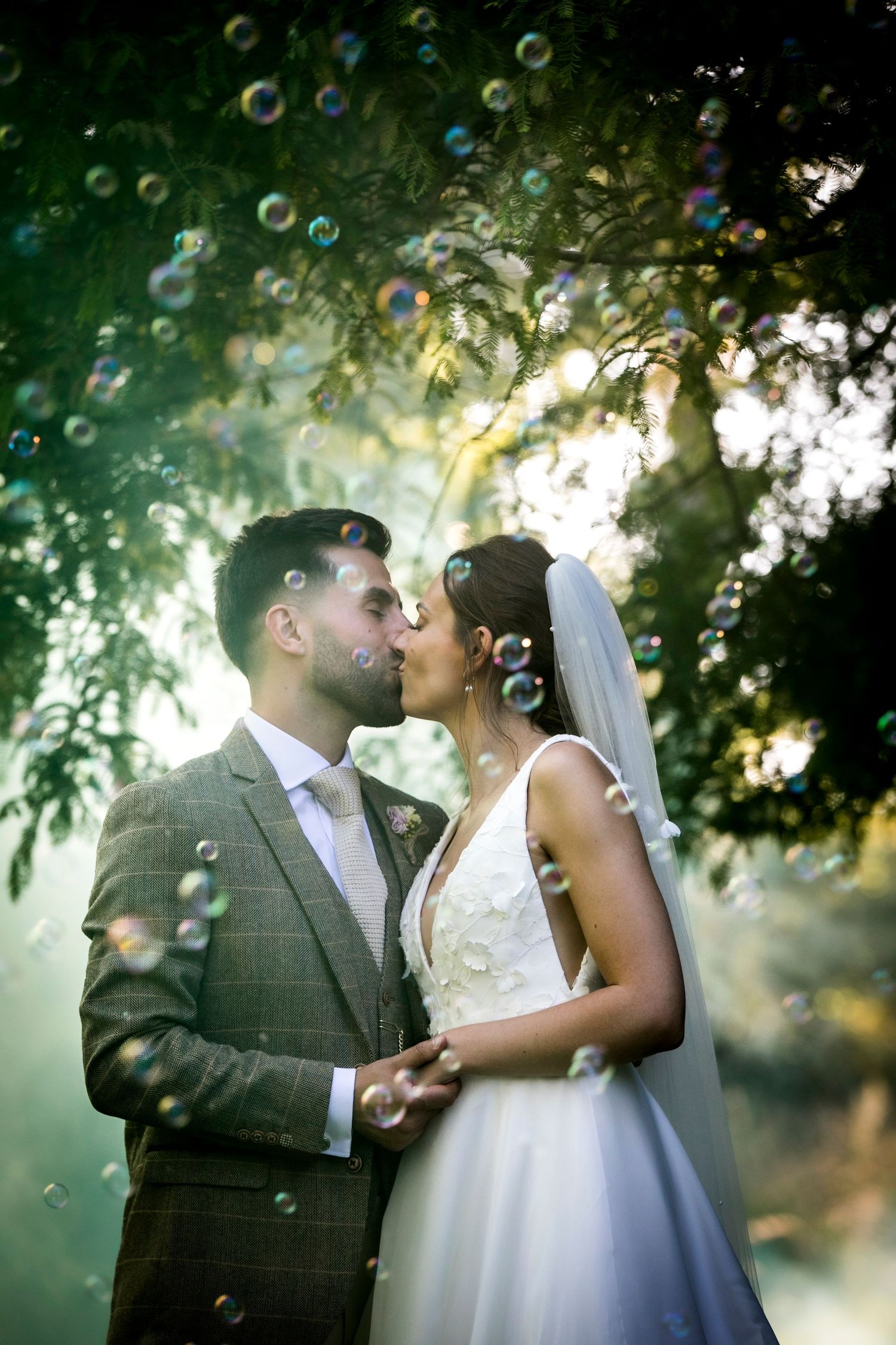 A romantic wedding couple shares a tender kiss outdoors with bubbles floating around them in soft natural lighting.