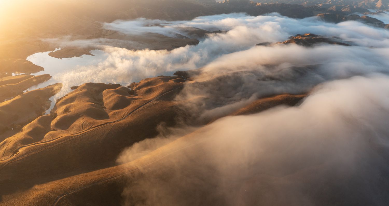 Aerial view of misty clouds rolling over golden mountain ridges at sunset casting dramatic shadows across the landscape.