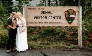 A couple poses in front of the Denali Visitor Center sign surrounded by evergreen forest.