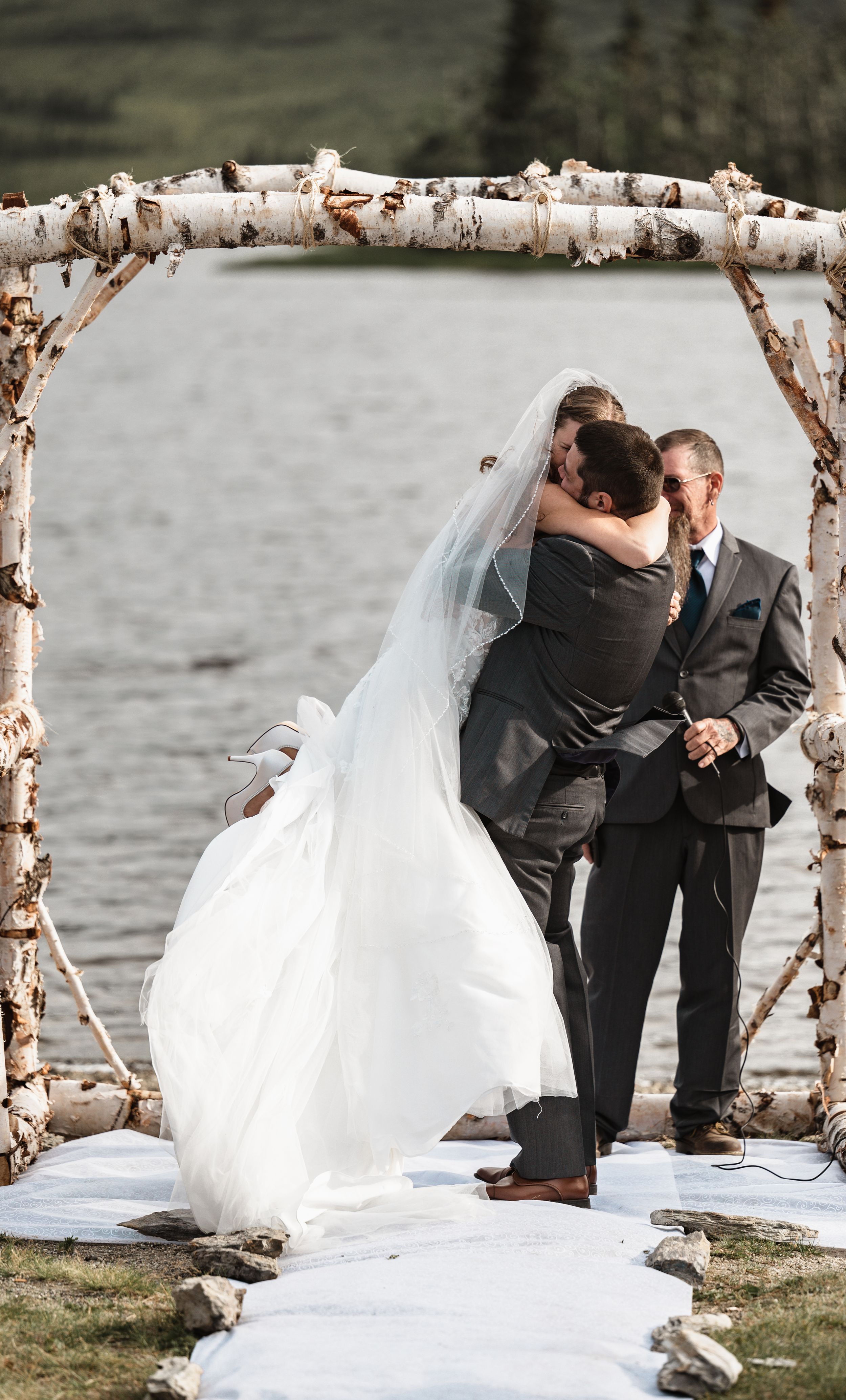 Wedding couple embraces under rustic birch wood arch on white aisle runner.