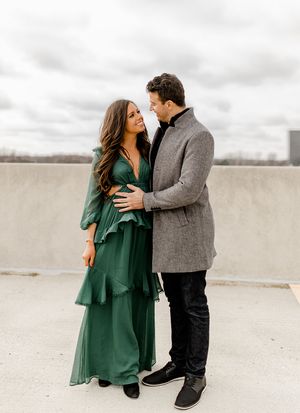 Romantic couple embracing outdoors, with woman in emerald dress and man in grey coat against cloudy sky.
