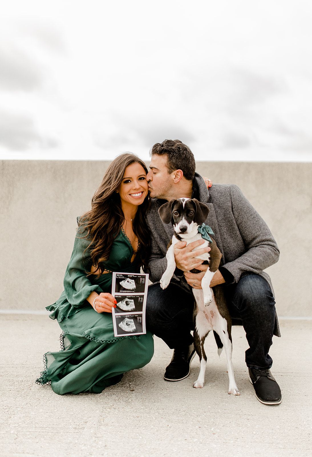 Couple kneeling with their dog while holding ultrasound photos in casual outdoor setting.