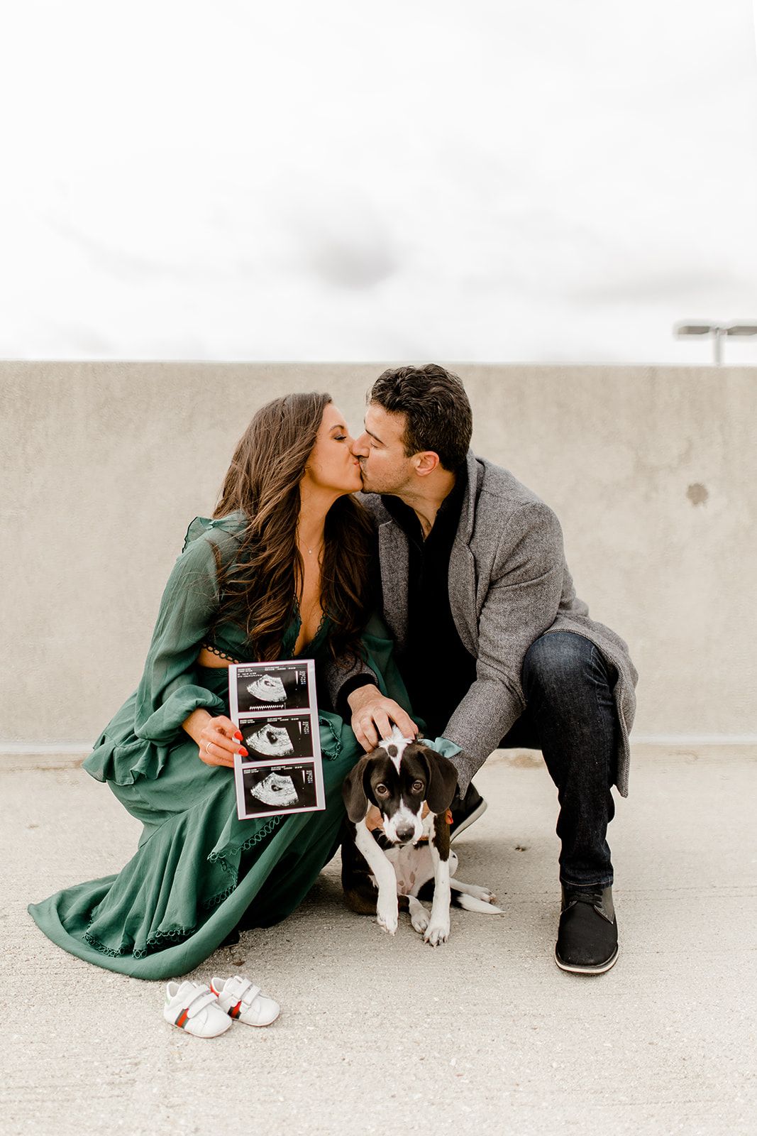 Couple kisses while holding ultrasound photos with their dog sitting beside them on concrete rooftop.