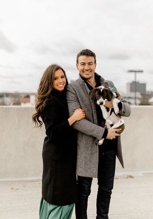 Happy couple poses on rooftop with their black and white dog while wearing winter coats.