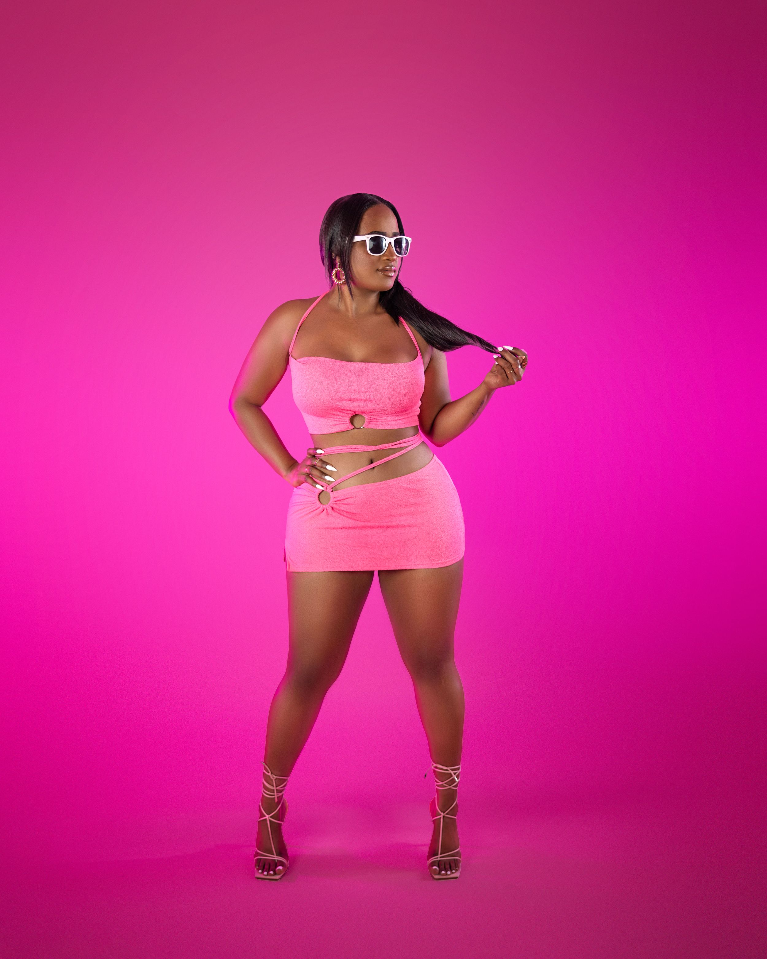 Model in pink two-piece outfit and sunglasses poses against bright pink background.