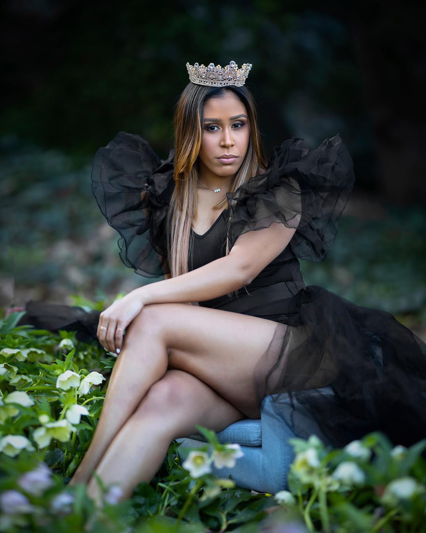 Model in black tulle dress and crown poses in garden setting with flowers.