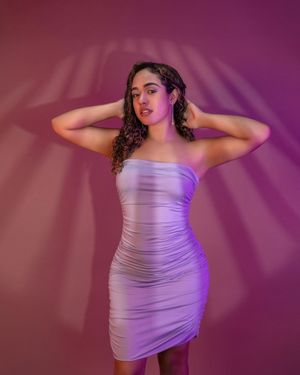 Fashion model in form-fitting lavender dress poses against pink backdrop with dramatic lighting.