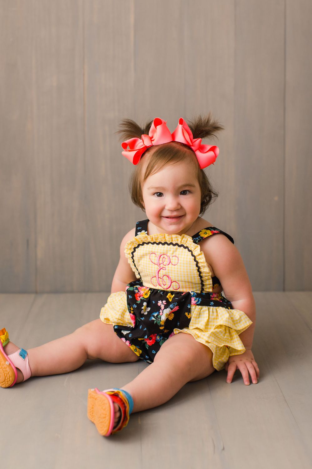 A toddler wearing a floral romper and red hair bows sits on a gray floor with colorful sandals.