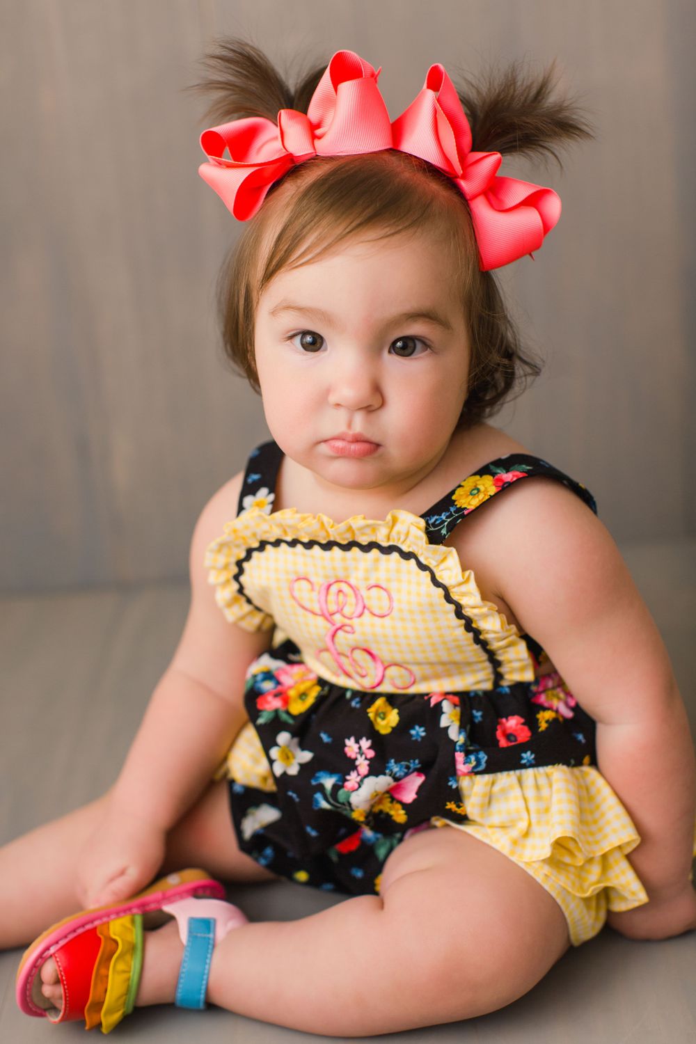 A small child in a yellow and navy floral outfit with pink bows sits on a studio floor.