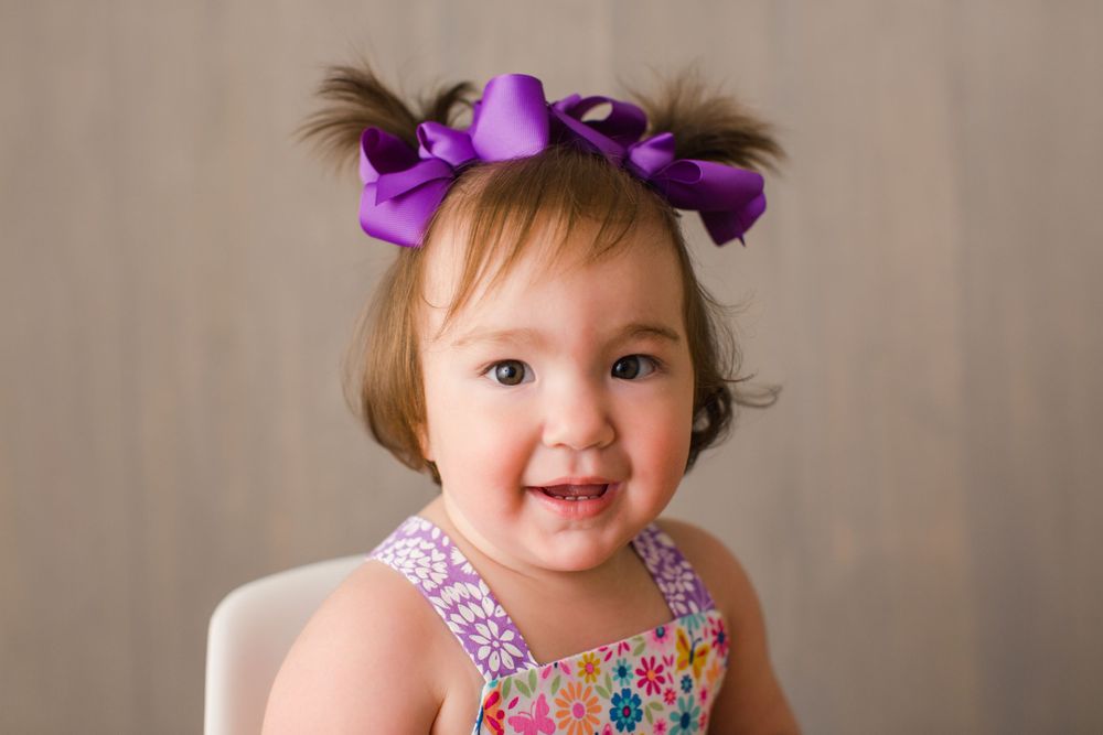 A smiling toddler wears a floral dress and purple hair bows against a neutral backdrop.
