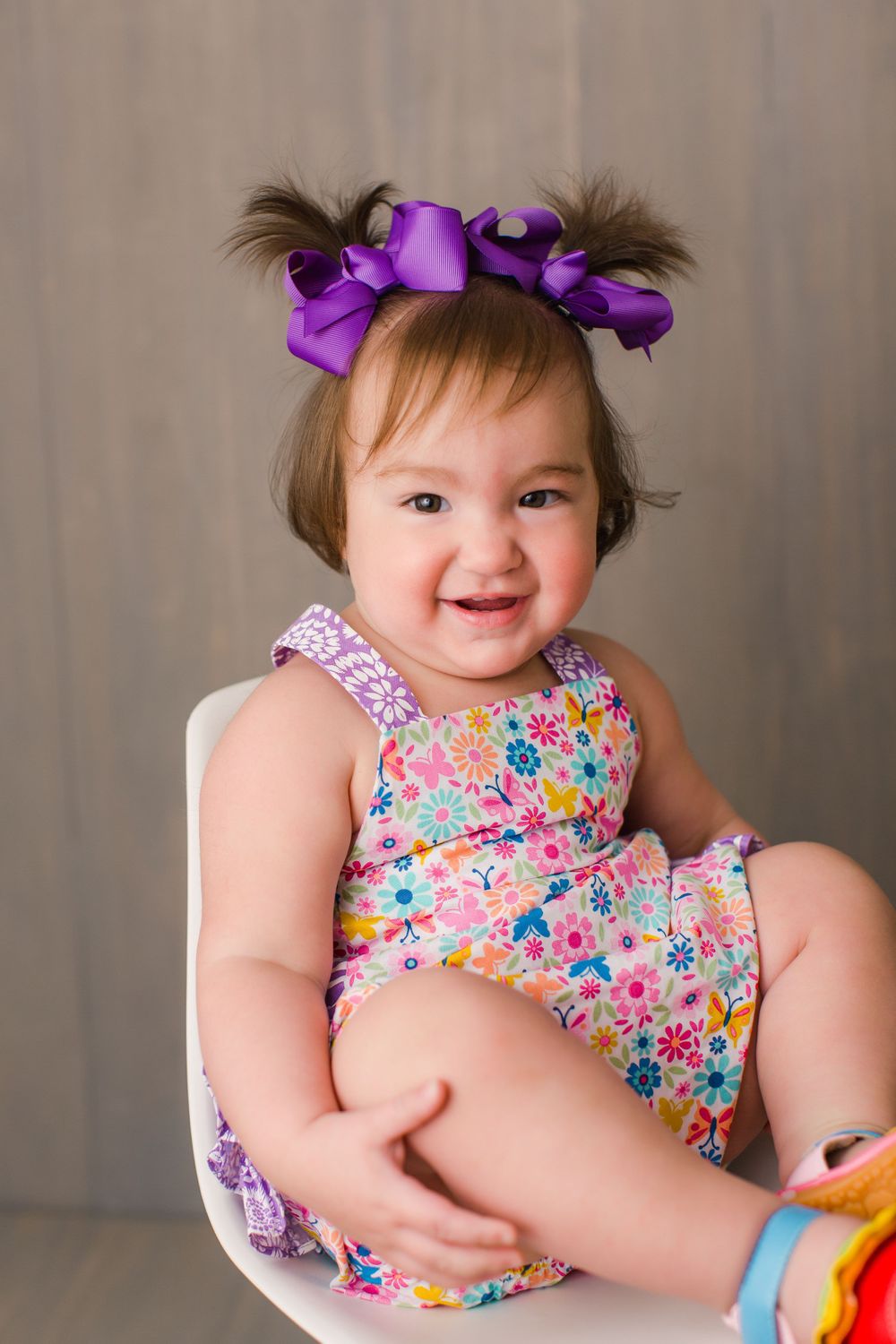A cheerful toddler in a colorful floral outfit and purple bows sits in a white chair.