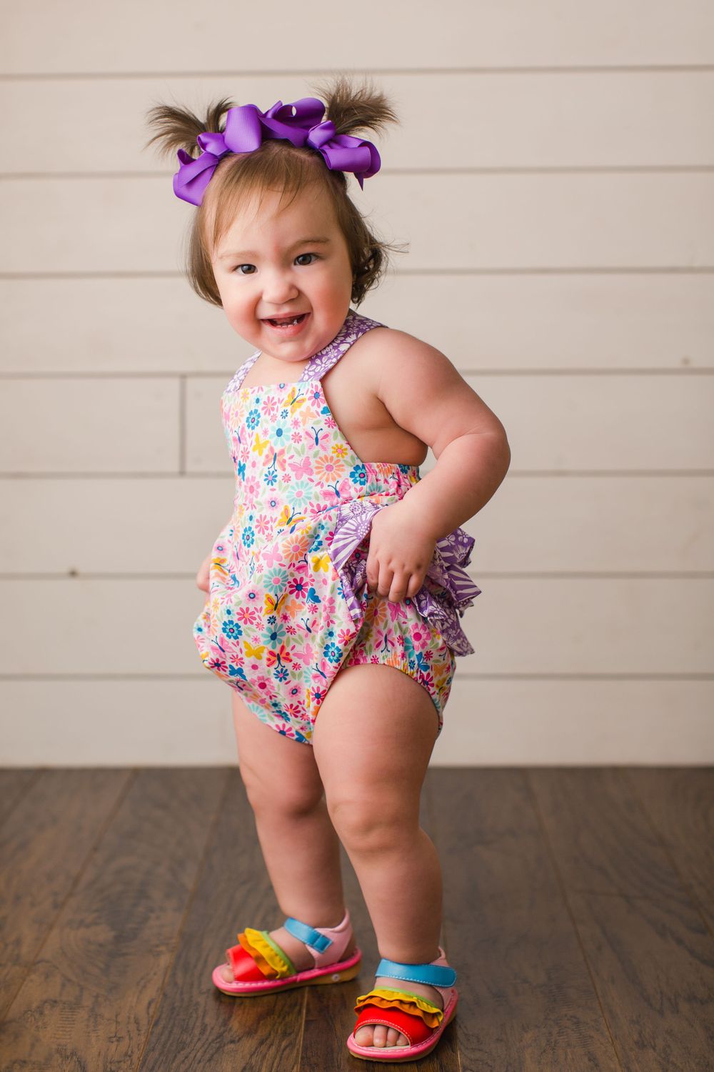 A joyful toddler in a floral romper and purple bows stands on hardwood flooring.