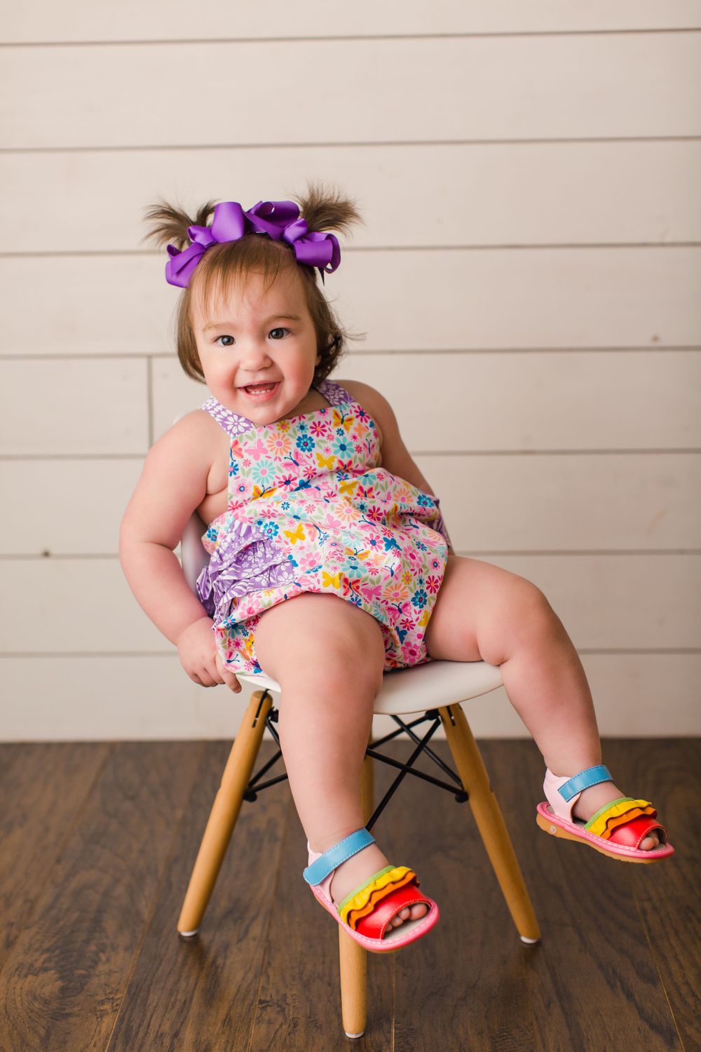 A toddler in a floral romper and purple bows sits on a modern chair against white planks.
