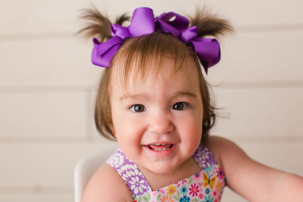 A happy toddler with purple hair bows wears a floral outfit in a studio setting.
