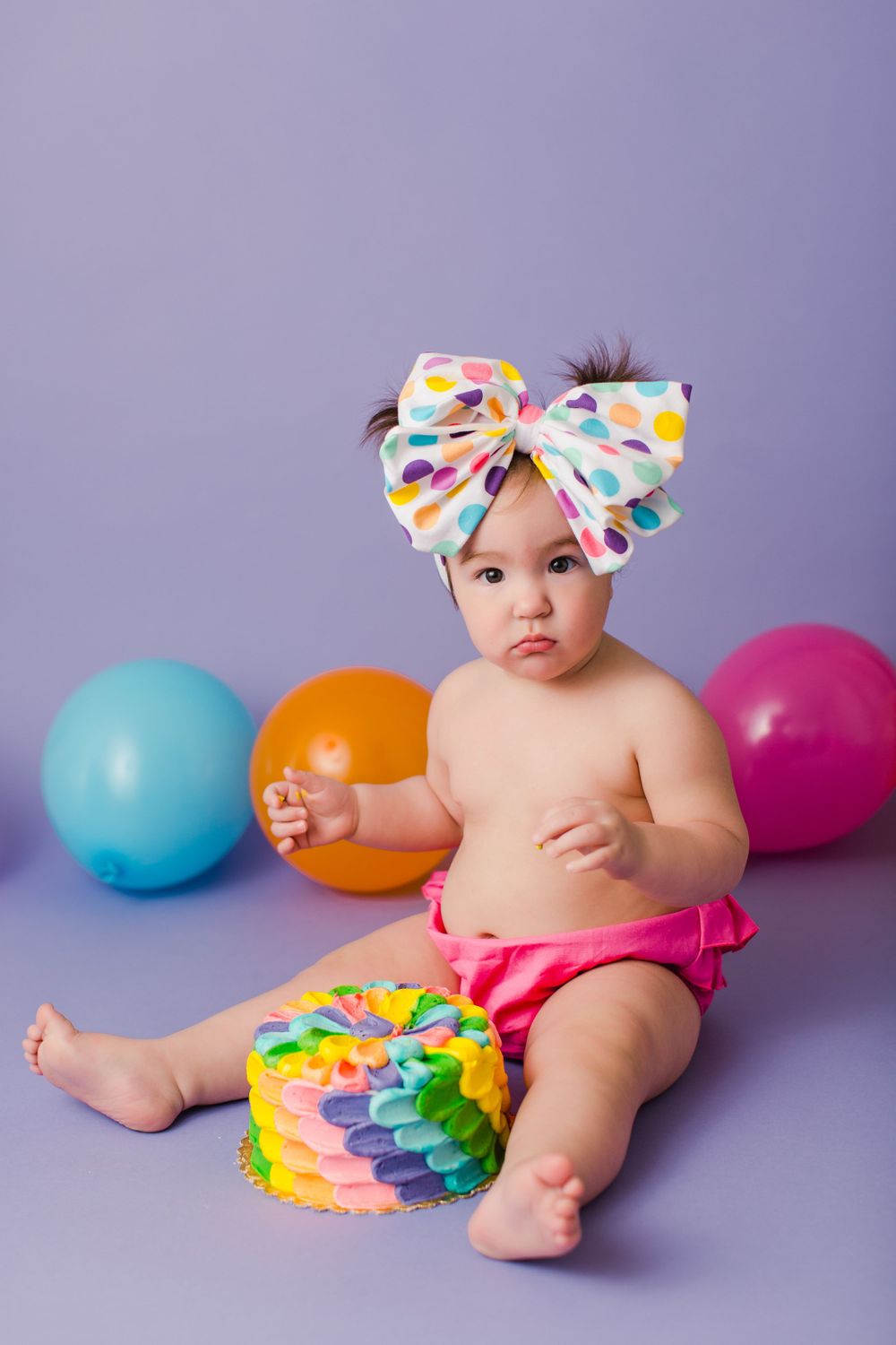 A baby wearing a colorful bow headband sits with balloons and a rainbow cake on purple backdrop.