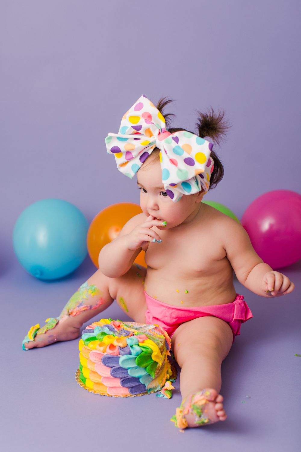 A baby in pink diaper and polka dot bow poses with colorful balloons and cake.