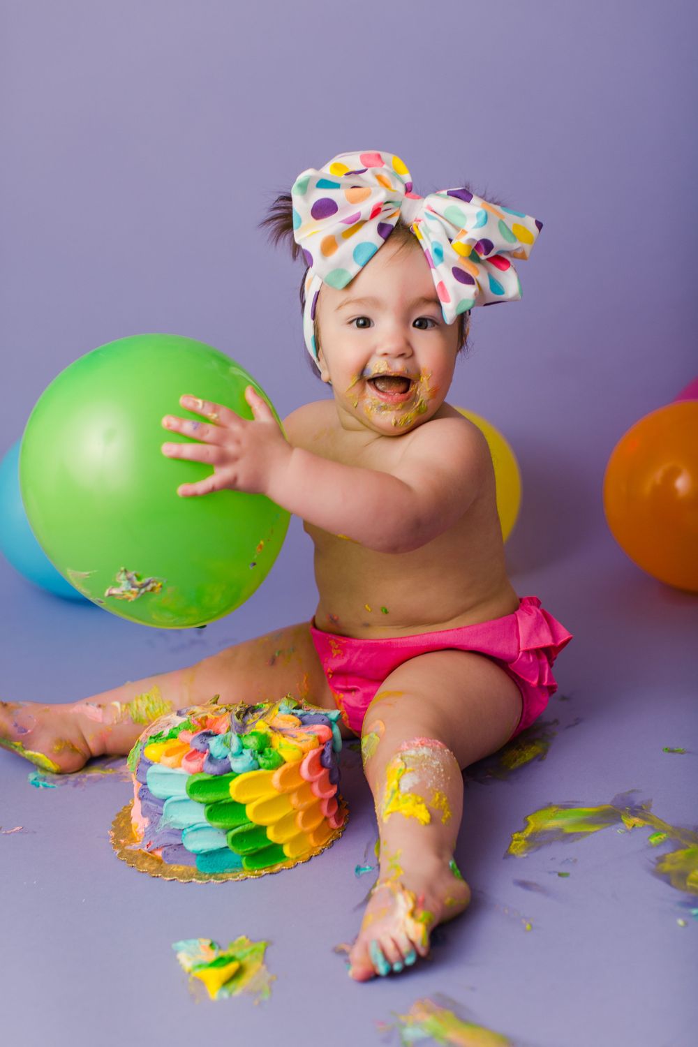 Smiling baby in polka dot bow and pink bloomers plays with colorful balloons and rainbow cake during messy cake smash photo shoot.