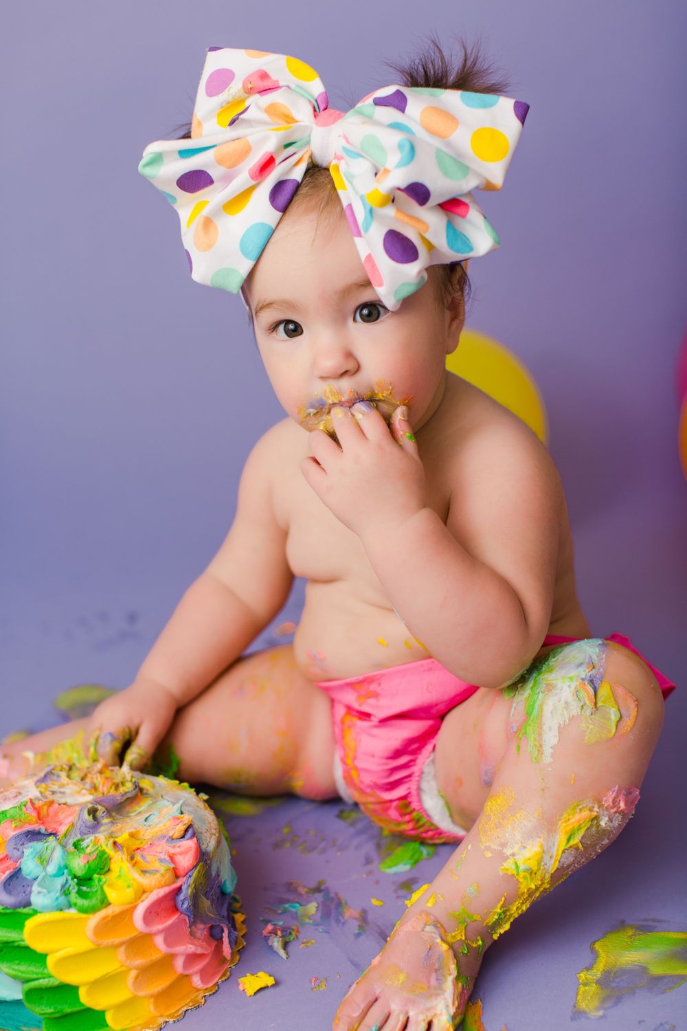 Baby wearing polka dot bow and pink diaper sits with colorful smashed cake and balloons against purple backdrop.