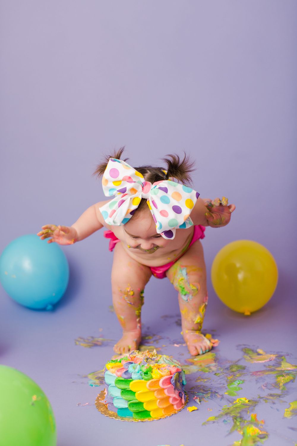 Baby in polka dot bow reaches for blue balloon during colorful cake smash session on purple background.